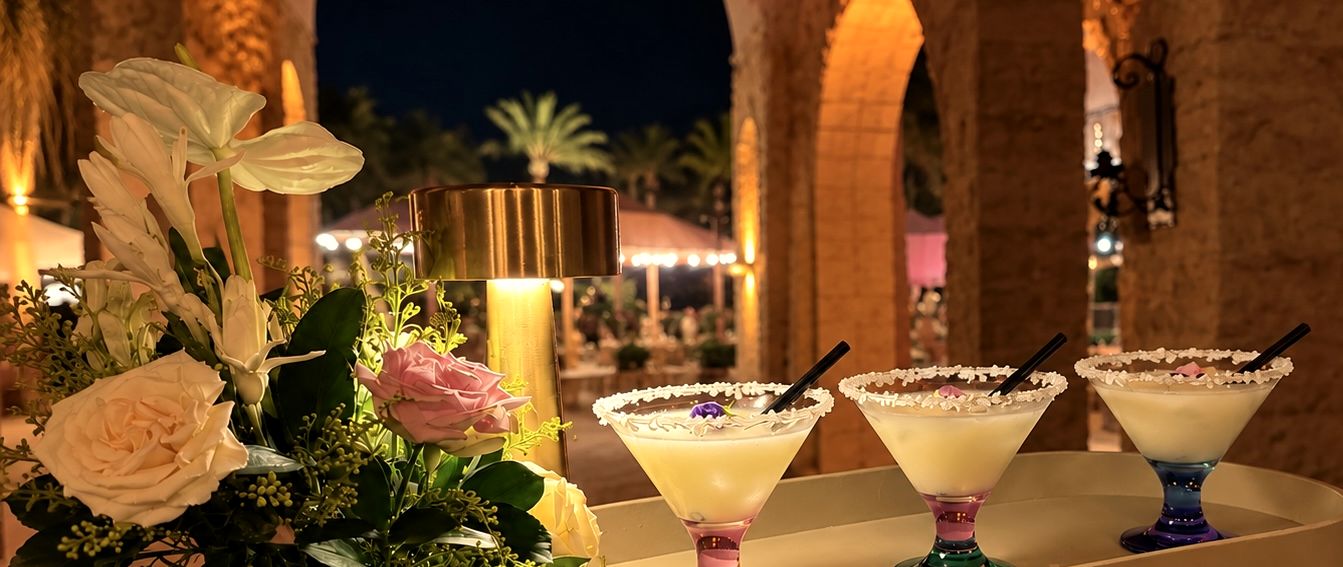 three sugar-rimmed drinks on a tray beside white flowers, under lit arches at night